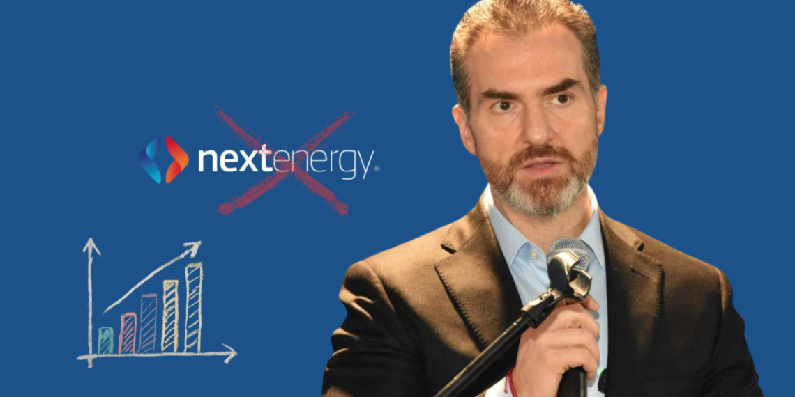 Man in a suit speaking into a microphone, with a chalk-drawn bar chart and a crossed-out NextEnergy logo on a blue background, conveying critique of the company’s growth claims.