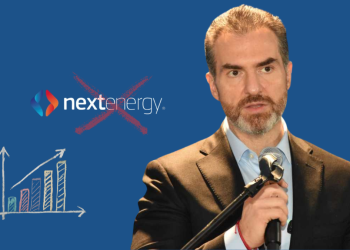 Man in a suit speaking into a microphone, with a chalk-drawn bar chart and a crossed-out NextEnergy logo on a blue background, conveying critique of the company’s growth claims.