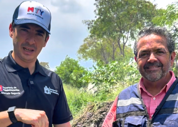 Two men pose outdoors in a sunny grassy area; one in a black polo with official logos and a cap, the other in a blue safety vest, smiling at the camera.