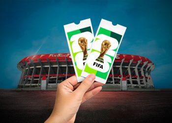 Hand holding two FIFA World Cup tickets in front of a large football stadium against a blue sky, suggesting event access.