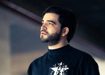 Portrait of a man with dark hair and beard, looking off to the side, wearing a black turtleneck-style shirt outdoors.